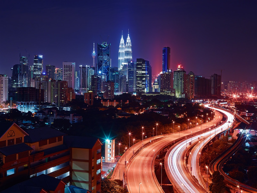 Nächtliche Stadtansicht von Kuala Lumpur mit den Petronas Towers und Lichtspuren von Fahrzeugen.